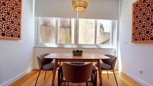 Uma sala de jantar com uma mesa de madeira, quatro cadeiras, um lustre e uma janela com vista para edifícios.