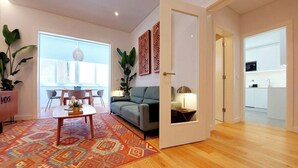 Uma sala de estar moderna com um sofá, mesa de centro e um tapete estampado. Há uma área de jantar com cadeiras e uma mesa. Uma cozinha é visível através de uma porta aberta.