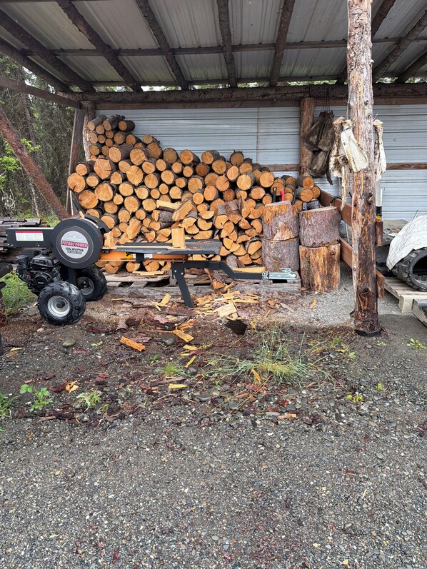 A covered outdoor area with a stack of logs, a log splitter, and a vehicle.