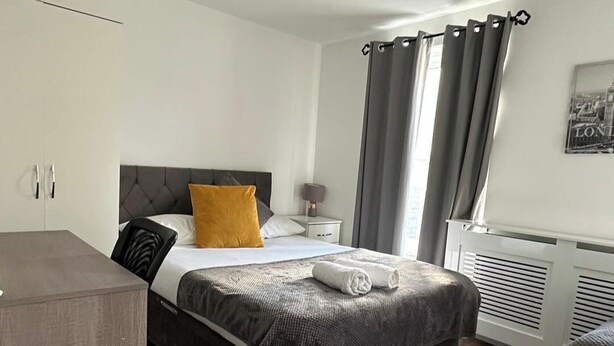 A hotel room with a bed, a bedside table, a lamp, a radiator, and a window with curtains. - House Sleeps 22 with Wifi (London)