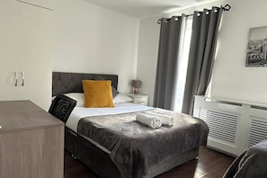 Quarto de hotel com cama, mesa de cabeceira, luminária, radiador e janela com cortinas.