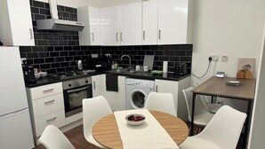 A compact kitchen with white cabinets, a black tiled backsplash, a stove, an oven, a washing machine, and a small dining table with chairs. - House Sleeps 22 with Wifi (London)
