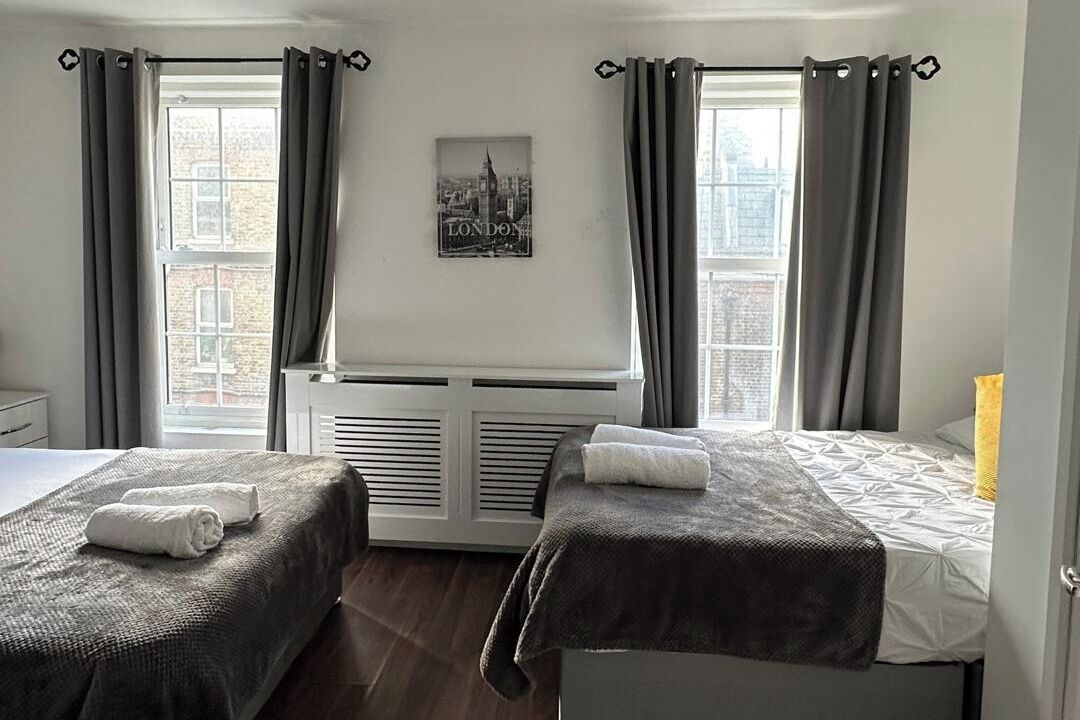 Ein Hotelzimmer mit zwei Betten, einem Heizkörper, einem Stadtbild an der Wand und einem Fenster mit Vorhängen.