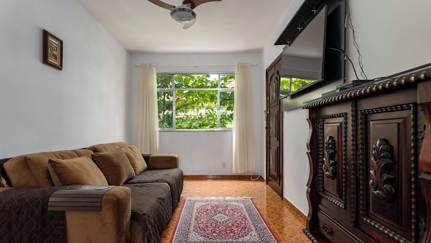 A room with a sofa, a wooden cabinet, a television, a window with curtains, and a patterned rug. - RJ JACAREPAGUÁ - Cozy and spacious apartment. We accept small pets. (Rio de Janeiro)