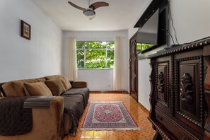 Ein Zimmer mit einer Couch, einem Holzschrank, einem Fernseher, einem Fenster mit Vorhängen und einem gemusterten Teppich.