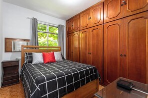 A bedroom with a bed, wooden wardrobe, a nightstand, a window with curtains, and a desk with a lamp. - RJ JACAREPAGUÁ - Cozy and spacious apartment. We accept small pets. (Arraial do Cabo)