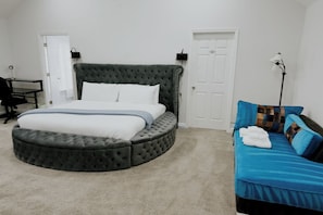 Une chambre à coucher avec un lit doté d’une tête de lit capitonnée, un pouf rond, un canapé bleu avec des coussins à motifs, un bureau et une lampe sur pied.