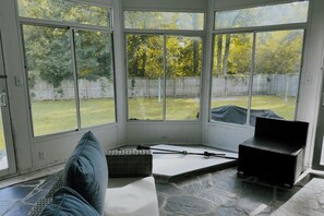 Ein Zimmer mit großen Fenstern, Steinboden und Blick auf einen Hinterhof mit Holzzaun und Bäumen.