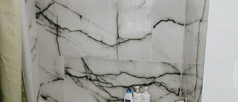 A bathroom with a marble wall, a bathtub, and toiletries on the edge.
