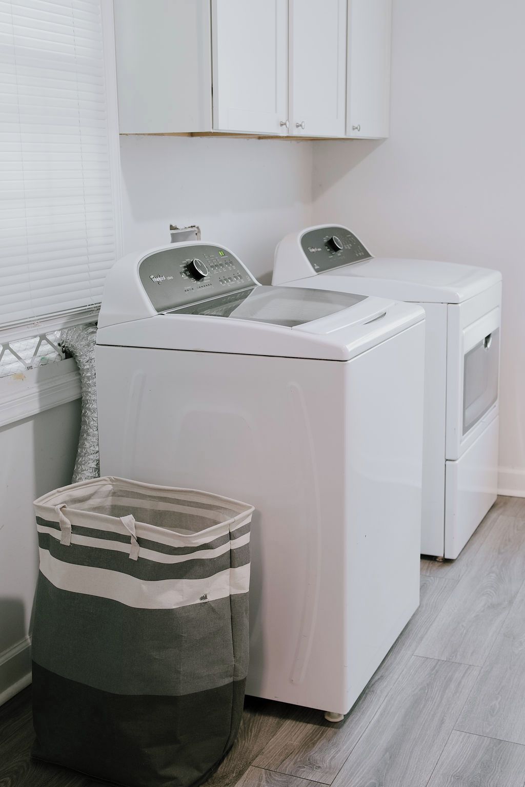 A laundry room with a front-loading washer and dryer, a striped laundry basket, and white cabinets above.