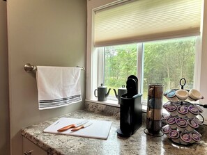 Una cocina con ventana, una toalla colgando en la puerta, una tabla de cortar con cuchillos, una cafetera y un escurreplatos.