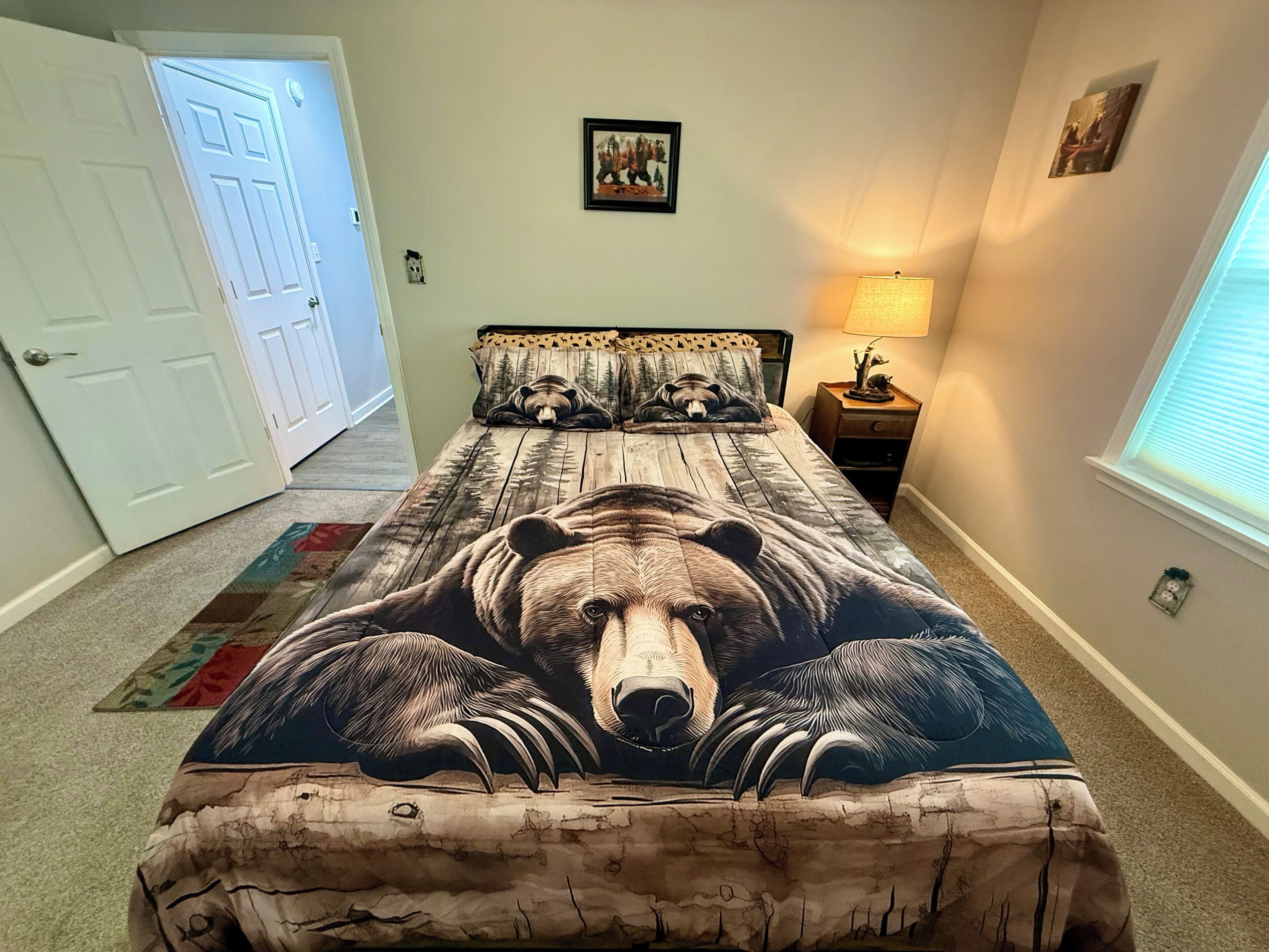 A bedroom with a bed featuring a bear-themed bedspread, a door, a nightstand with a lamp, and a window with blinds.