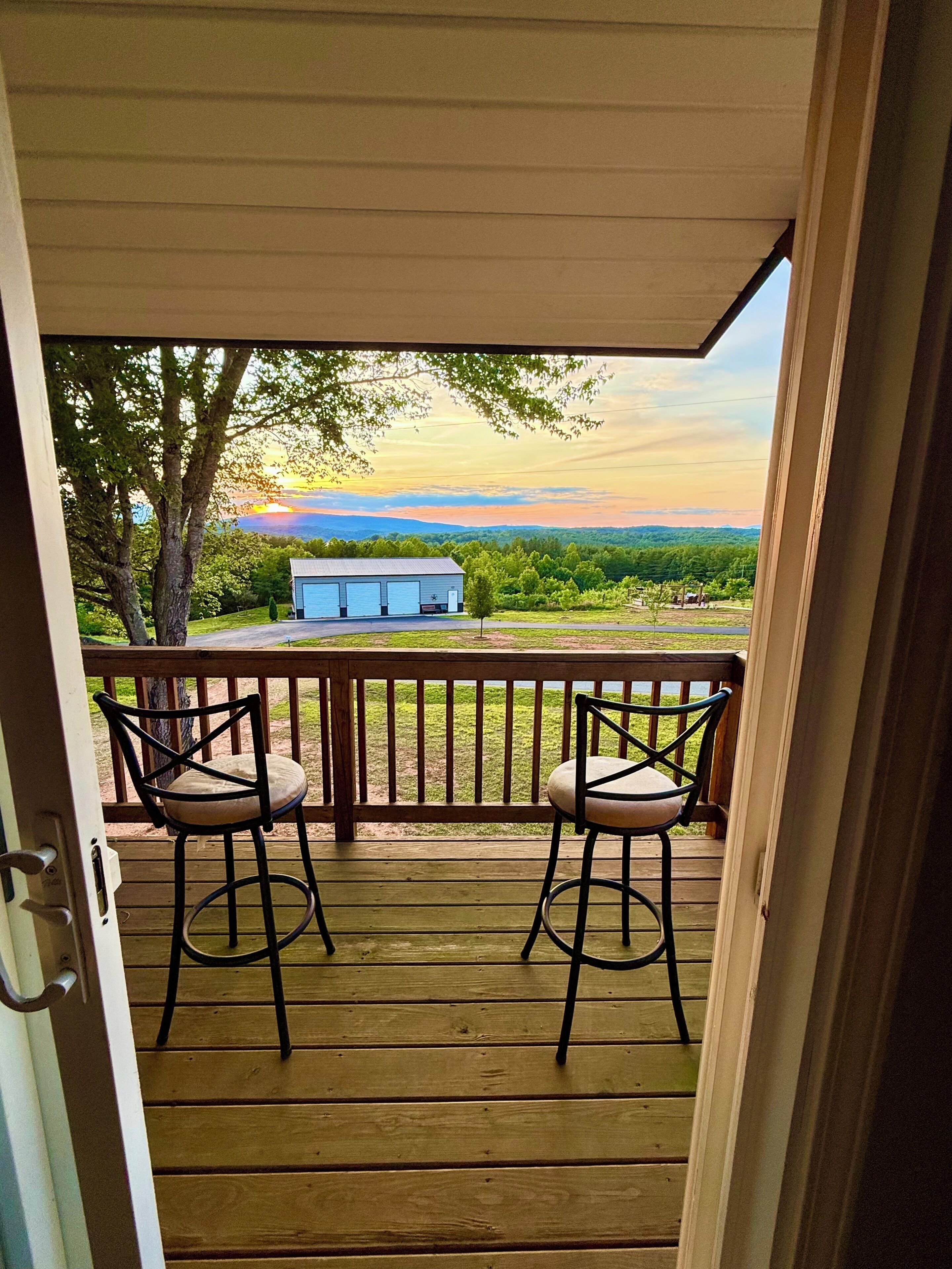 Une porte ouverte donne sur une terrasse en bois, où se trouvent deux chaises, et offre une vue sur un bâtiment et un paysage.