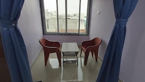 Ein Zimmer mit einem kleinen Tisch, zwei Stühlen, einem Fenster mit Blick auf Gebäude und blauen Vorhängen.