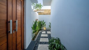Un sendero estrecho que lleva a una puerta de madera con manijas metálicas, rodeado de macetas con plantas y piedras para pisar.
