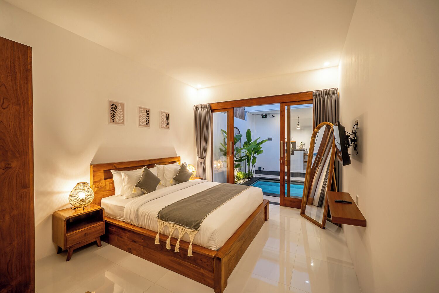 A bedroom with a wooden bed, a nightstand, a bedside lamp, a large mirror, and a sliding glass door leading to a pool area.