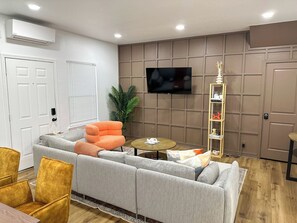 Ein Wohnzimmer mit grauem Sofa, einem orangefarbenen Sessel, einem kleinen runden Tisch, einem an der Wand montierten Fernseher und Holzboden.