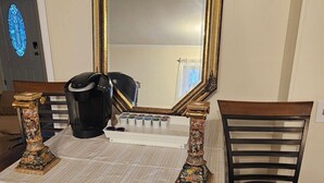 Taula de dinar amb mantell blanc, cadires ornamentals, un mirall gran amb marc daurat i una cafetera.