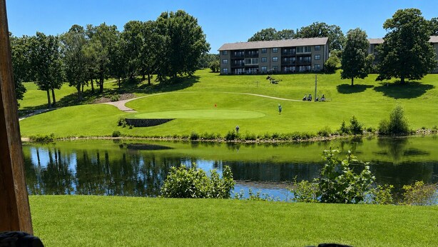 A view of a golf course with a pond, a building in the background, and wicker chairs in the foreground. - Relax & Unwind~Cozy 1bed/1bath Condo with Beautiful Pond & Golf Views (Branson)