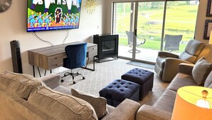 A living room with a TV, sofa, ottoman, and a view of a golf course through the glass door. - Relax & Unwind~Cozy 1bed/1bath Condo with Beautiful Pond & Golf Views (Branson)