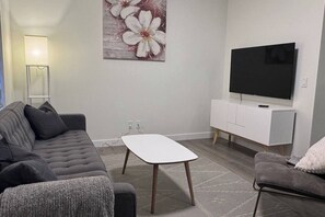 A living room with a grey sofa, a white coffee table, a flat-screen TV, and a white cabinet. - Heartland Nest - Brand New 2BR house with Garage, AC, BBQ (Cochrane)