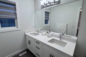 A bathroom with two sinks, a large mirror, and a window with blinds. - Heartland Nest - Brand New 2BR house with Garage, AC, BBQ (Cochrane)