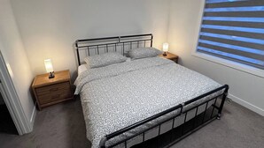 A single bed with a metal frame, two bedside tables with lamps, a window with blinds, and a carpeted floor. - Heartland Nest - Brand New 2BR house with Garage, AC, BBQ (Cochrane)