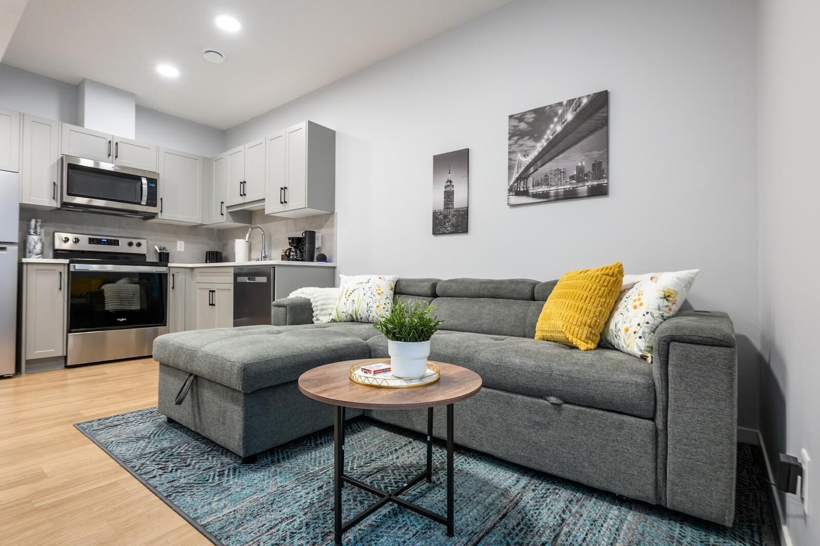 Een moderne woonkamer met een grijze bank, een klein rond tafeltje met een plant, en een keuken op de achtergrond.