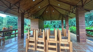 Un espacio al aire libre techado, con bancos de madera y suelo de baldosas, rodeado de vegetación.