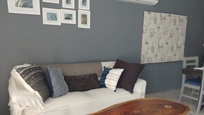 Una habitación con un sofá blanco, una mesa de centro de madera, una pared gris con cuadros enmarcados, un aire acondicionado y una cortina estampada.