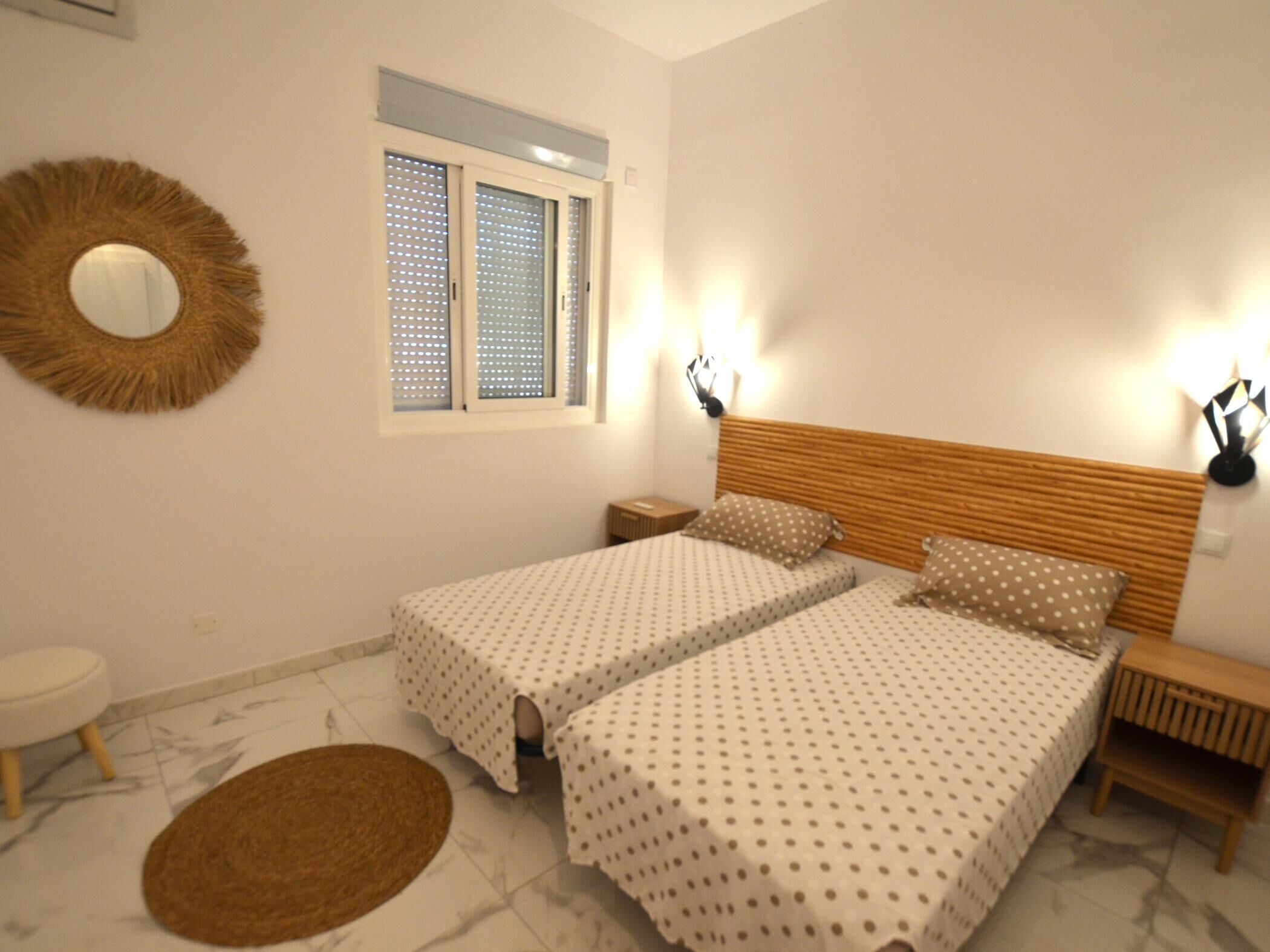 A bedroom with two beds, a round mirror, a window with blinds, and a nightstand.