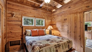 A wooden cabin bedroom with a bed, two windows, a ceiling fan, and a nightstand.