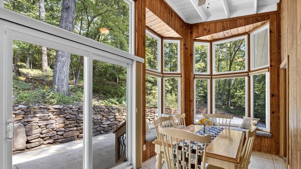 A wooden interior with a stone wall outside, a dining table with chairs, and large windows.