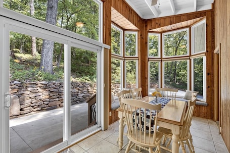 A wooden interior with a stone wall outside, a dining table with chairs, and large windows.
