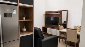 Een compacte woonruimte met een ingebouwde koelkast, een flatscreen televisie, een bank, een eettafel met stoelen en een kleine potplant.