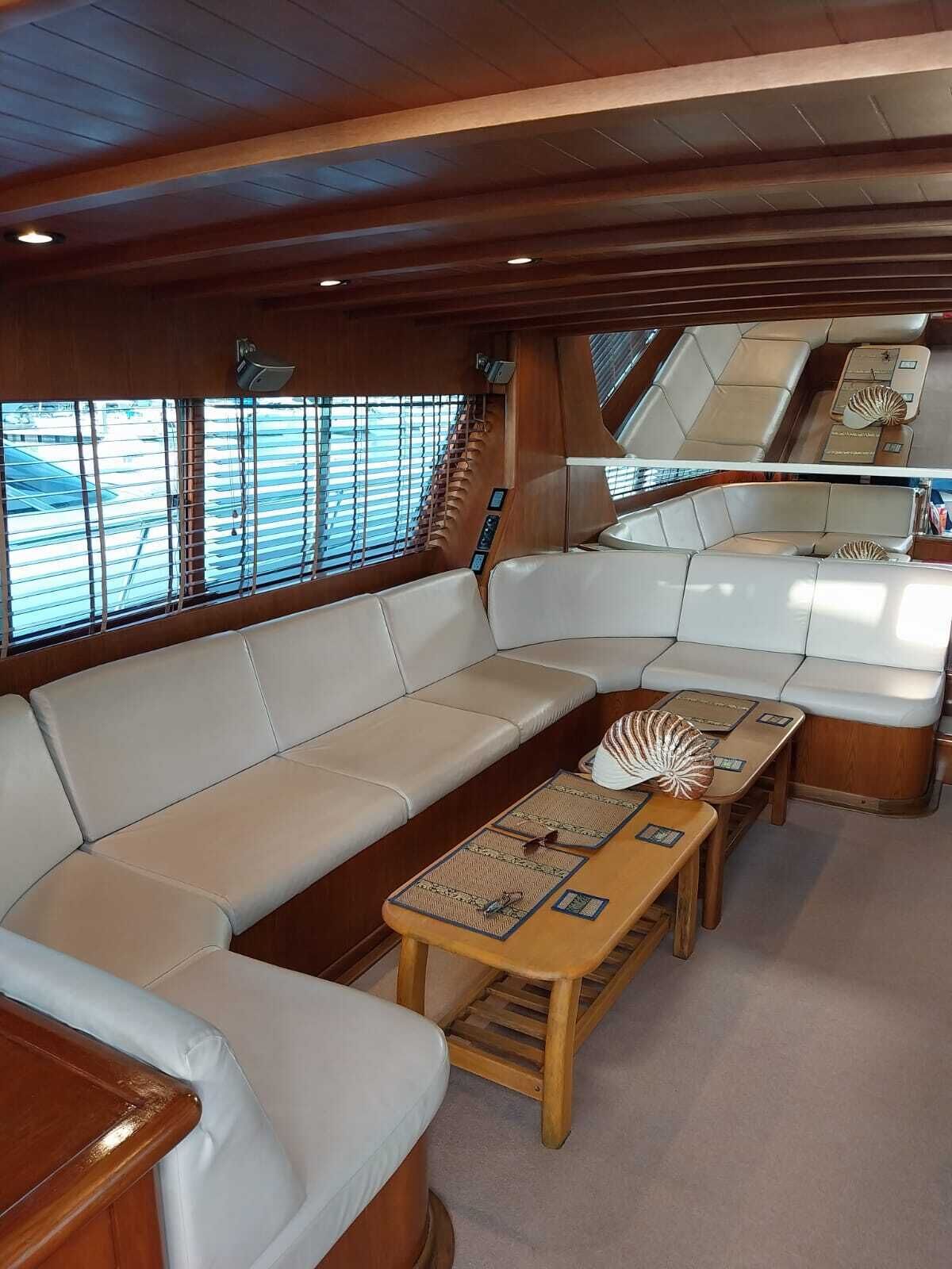 A yacht interior with a large cushioned seating area, a wooden table with a decorative shell, and a view of the marina through the windows.