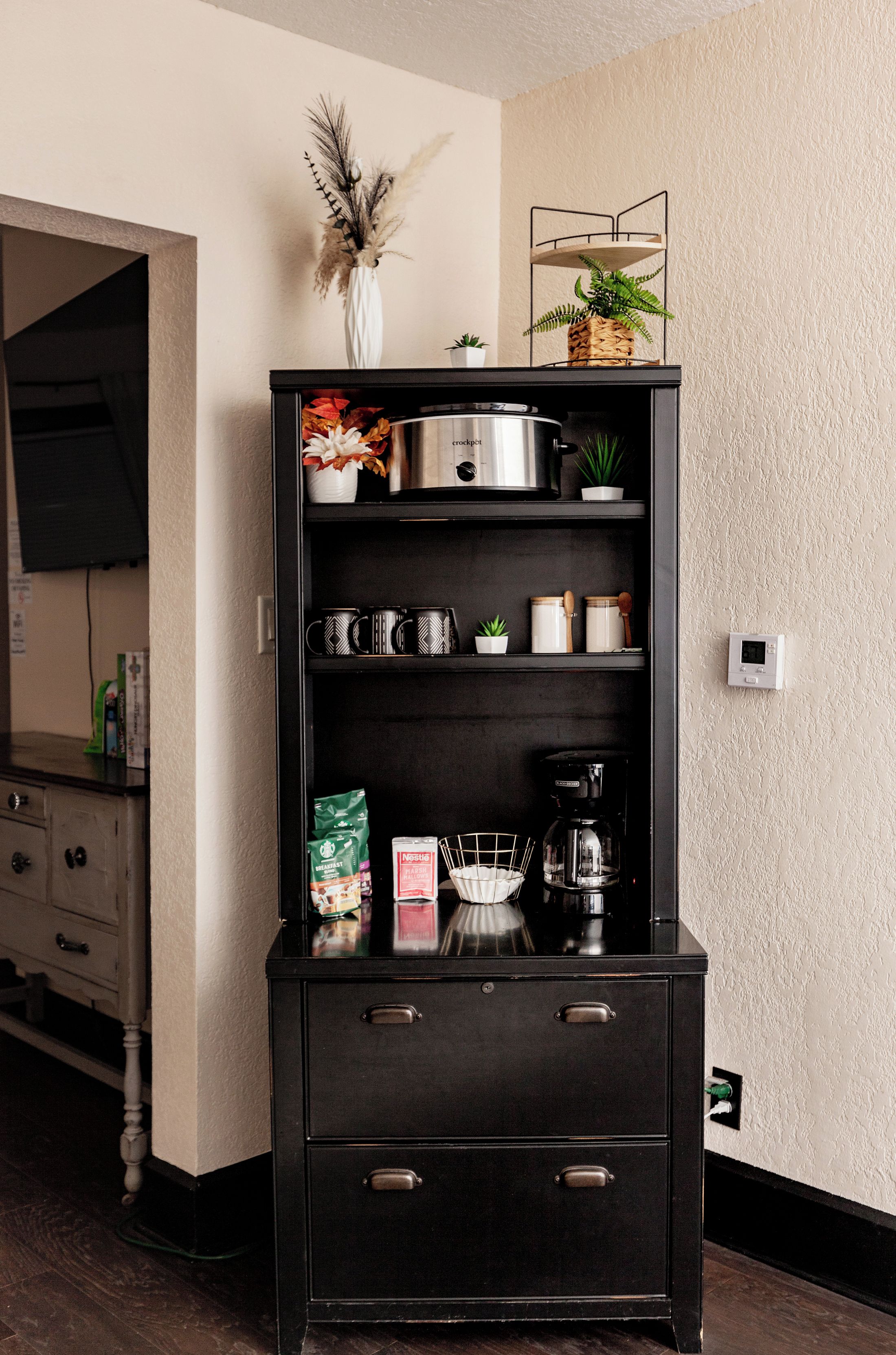 Eine Küche mit einem schwarzen Schrank, der eine Kaffeemaschine, einen Wasserkocher und eine Mikrowelle beherbergt.