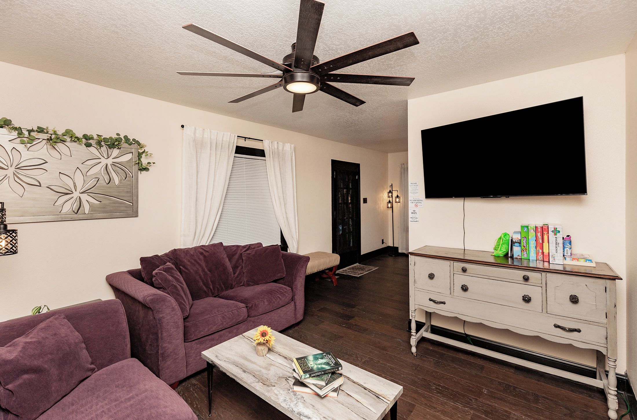 A living room with a purple sofa, a coffee table with books, a TV on the wall, and a wooden cabinet.