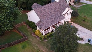 Une maison à deux étages dotée d’un porche couvert, entourée d’une cour clôturée et d’arbres.