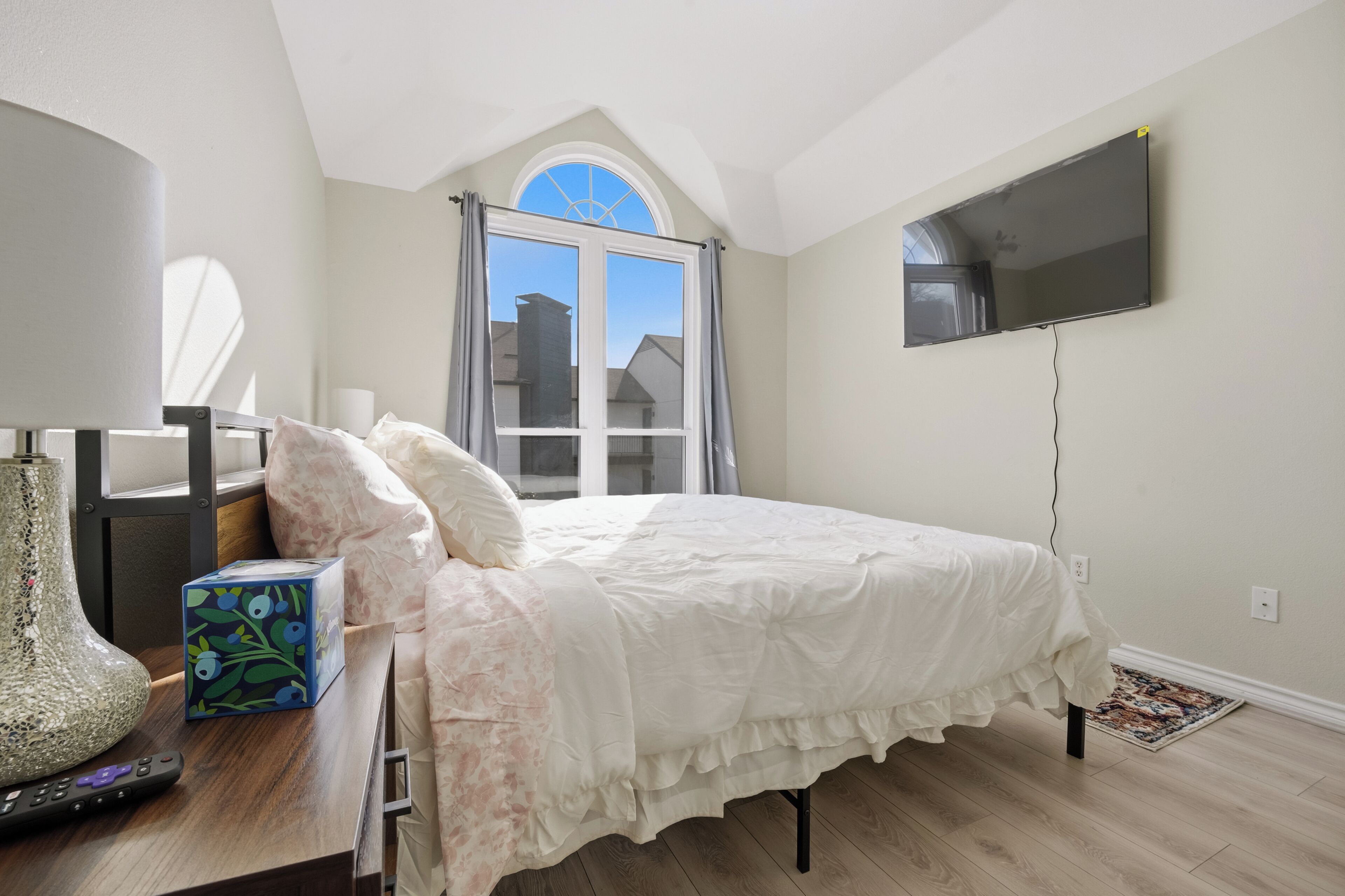 Une chambre à coucher avec un lit, un téléviseur à écran plat, une fenêtre avec des rideaux, un vase et une boîte posée sur une table de chevet.