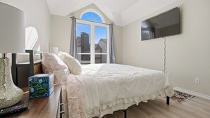 Une chambre à coucher avec un lit, un téléviseur à écran plat, une fenêtre avec des rideaux, un vase et une boîte posée sur une table de chevet.