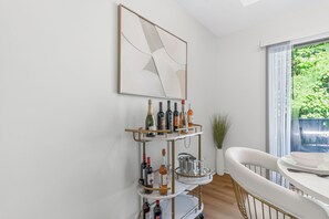 A modern kitchen with a white marble countertop, a gold and white metal cart with bottles, a large abstract painting, and a view of greenery outside.