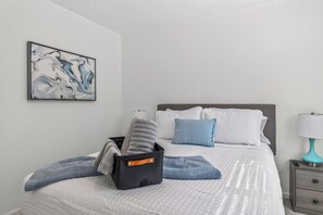 A neatly made bed with a quilted white cover, a grey pillow, and a blue pillow. A black bag with an orange strap is on the bed.