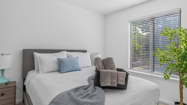En snyggt bäddad säng med kuddar, ett sängbord med en lampa, ett fönster med persienner och en krukväxt.