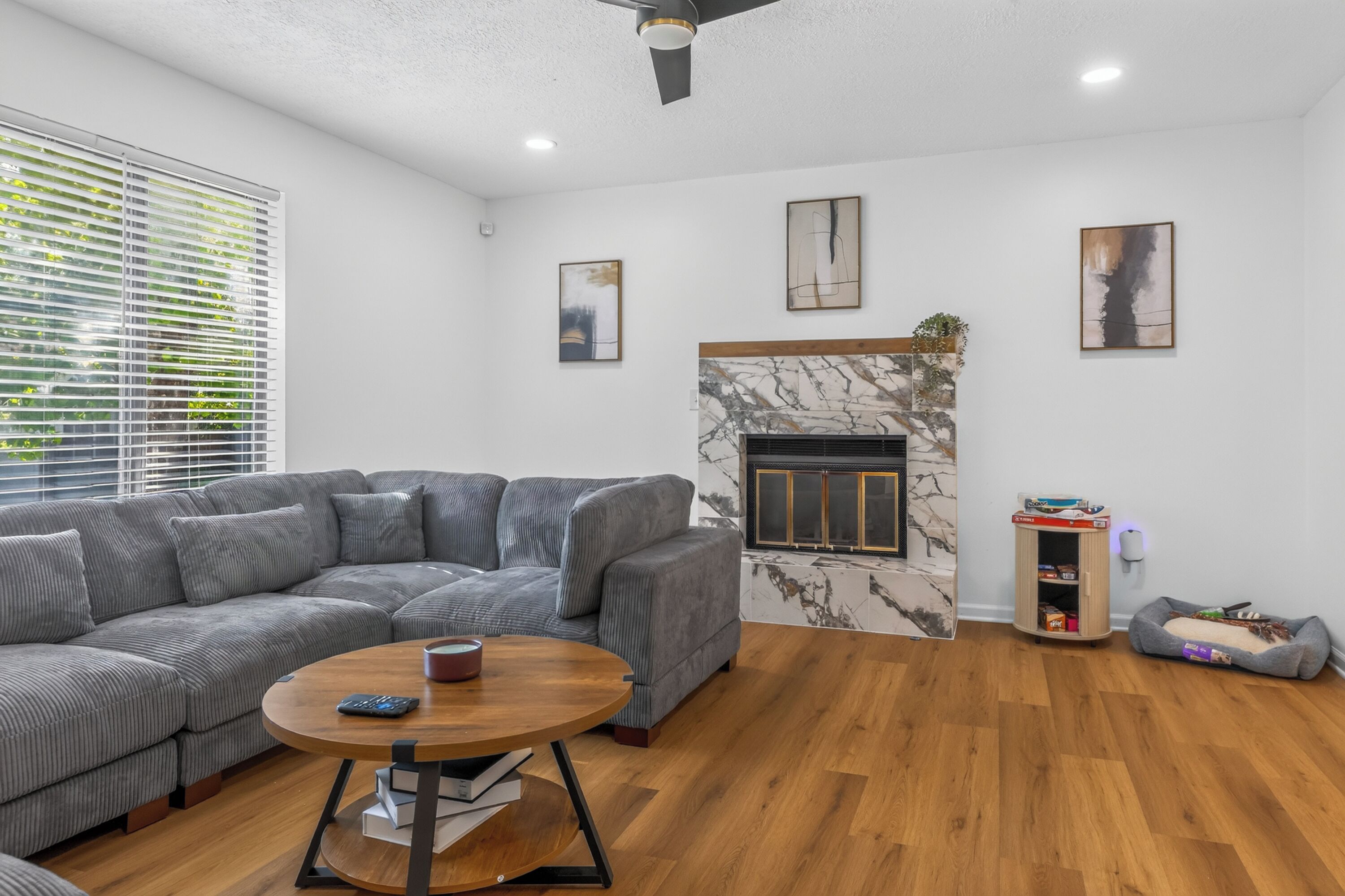 A modern living room with a grey sectional sofa, a round wooden coffee table, a fireplace with a marble surround, and a small wooden cabinet with a pet bed.