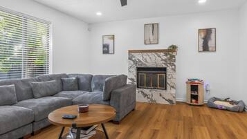 A modern living room with a grey sectional sofa, a round wooden coffee table, a fireplace with a marble surround, and a small wooden cabinet with a pet bed.