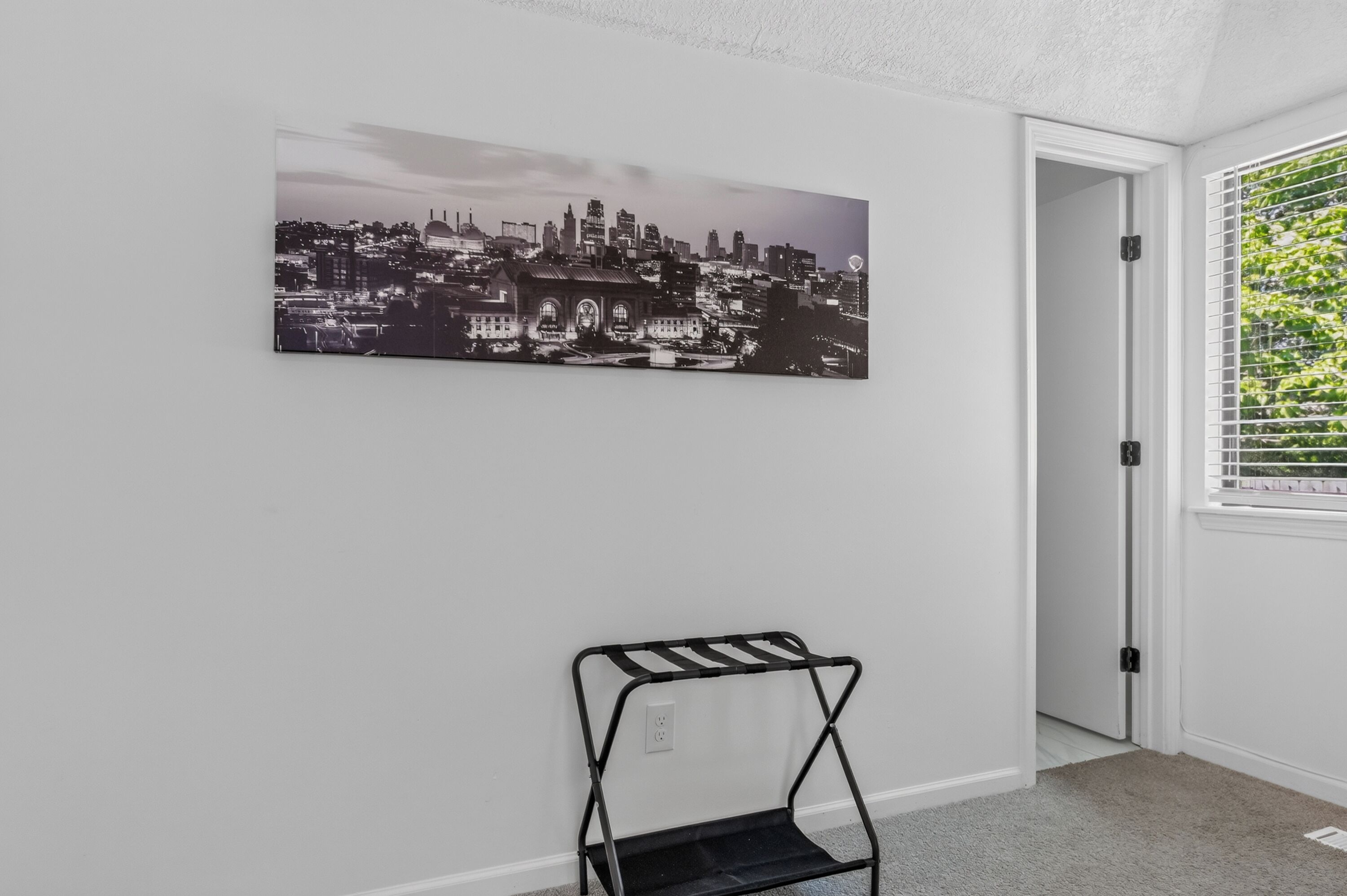 A room with a cityscape painting on the wall, a black metal shelf, and a door leading to a room with a window.