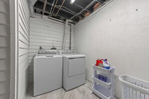A laundry room with two front-loading washing machines, a shelf with cleaning supplies, and a white laundry basket. - The Grand Estate . Private Pool . Sleeps 10 . Events Welcome (Grandview)