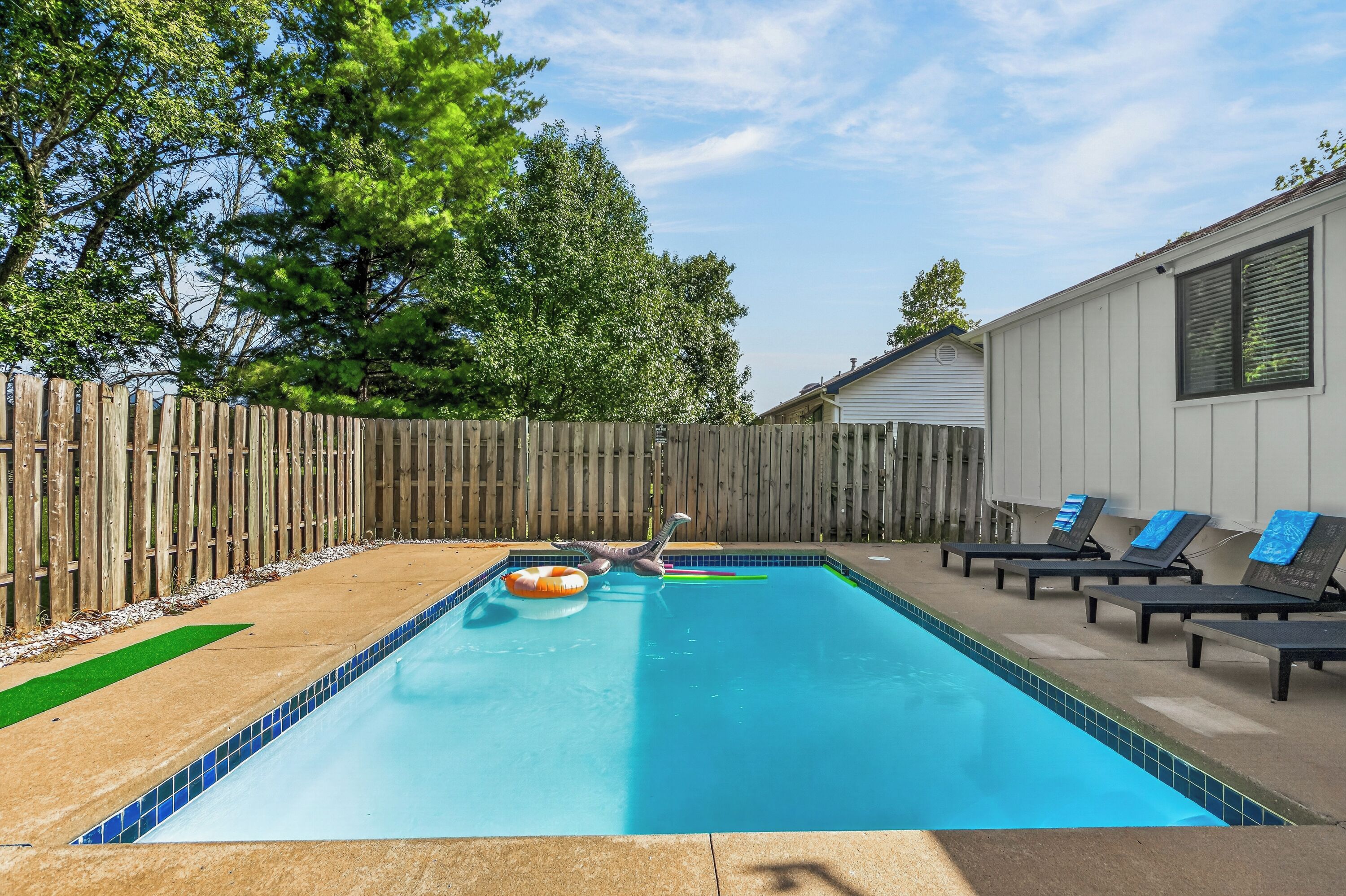 A rectangular swimming pool with a wooden fence and lounge chairs on one side.