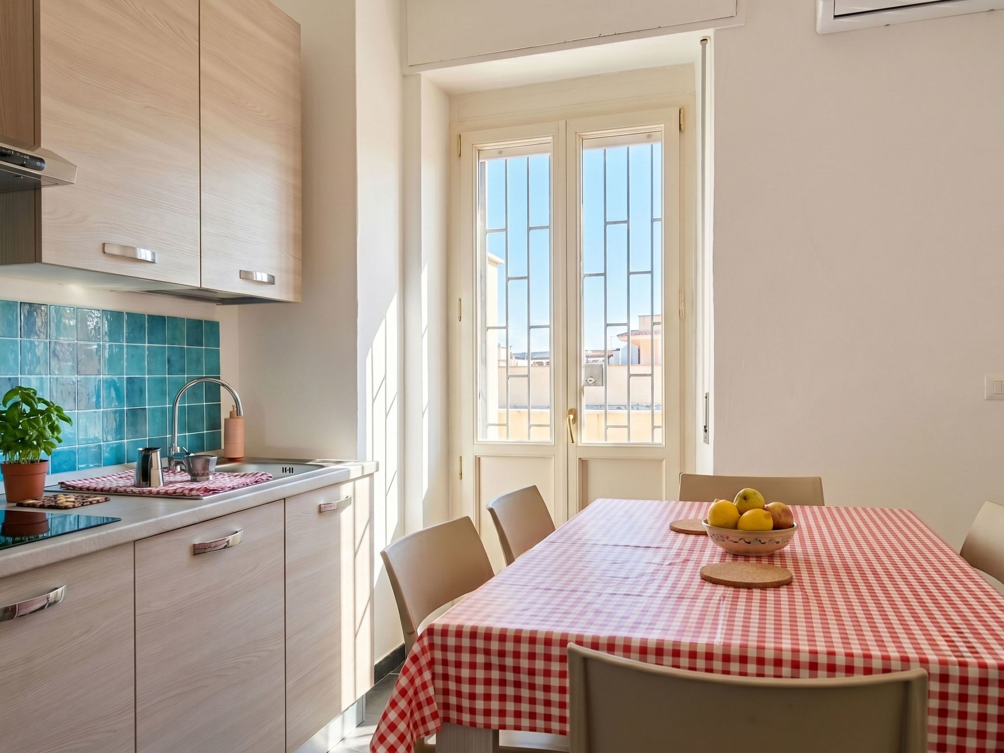 Una cucina con una finestra, un tavolo con tovaglia a quadri e una ciotola di frutta.